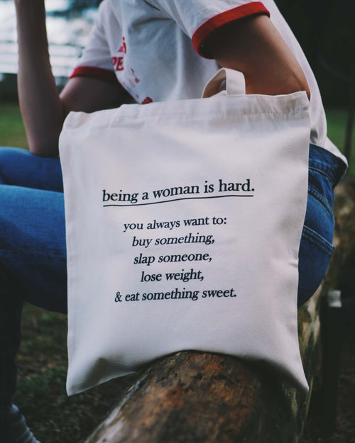 Tote bag with text about being a woman held by a person sitting outdoors.