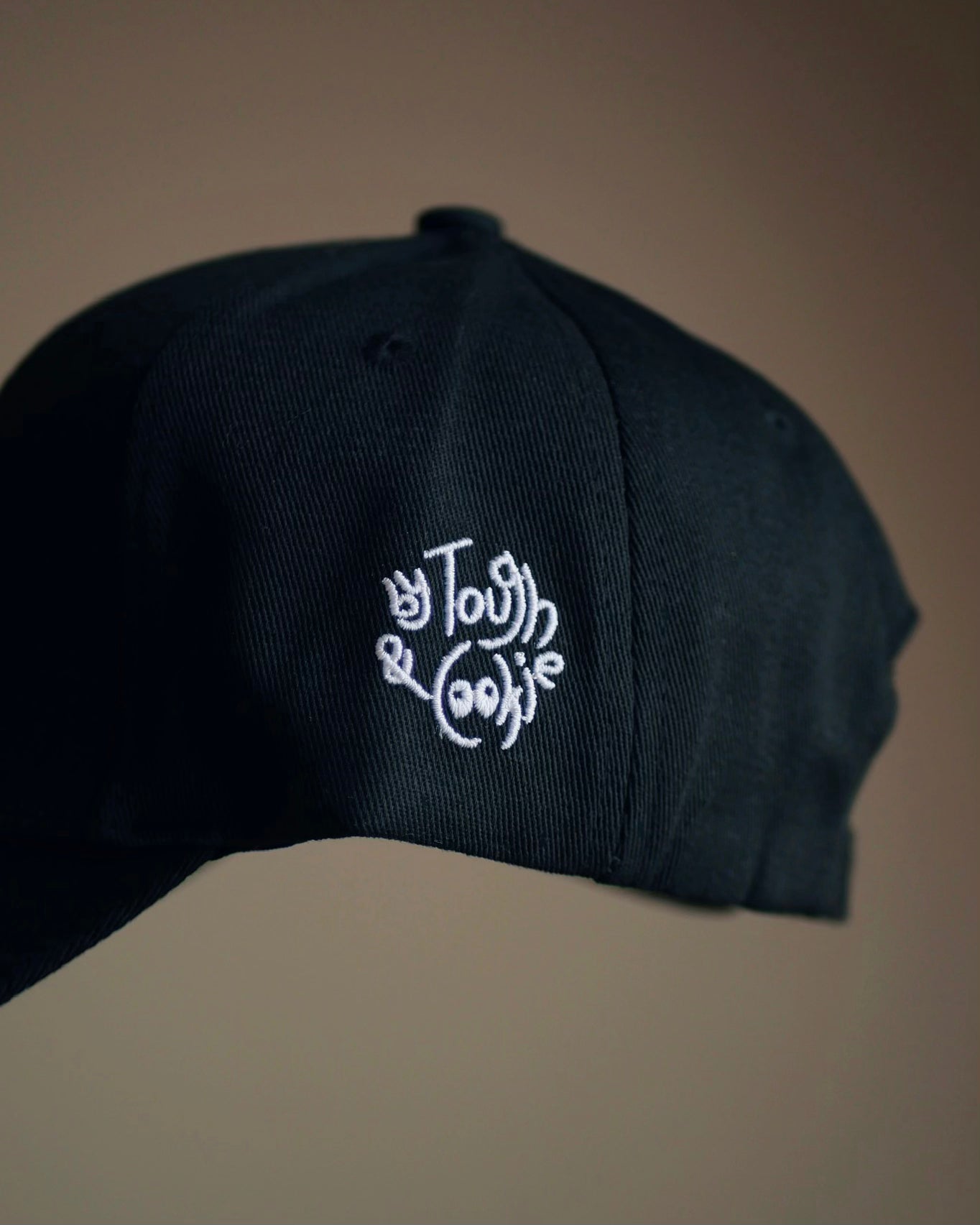 A close-up of a black baseball cap featuring white embroidered logo on the side panel, set against a solid warm brown background.