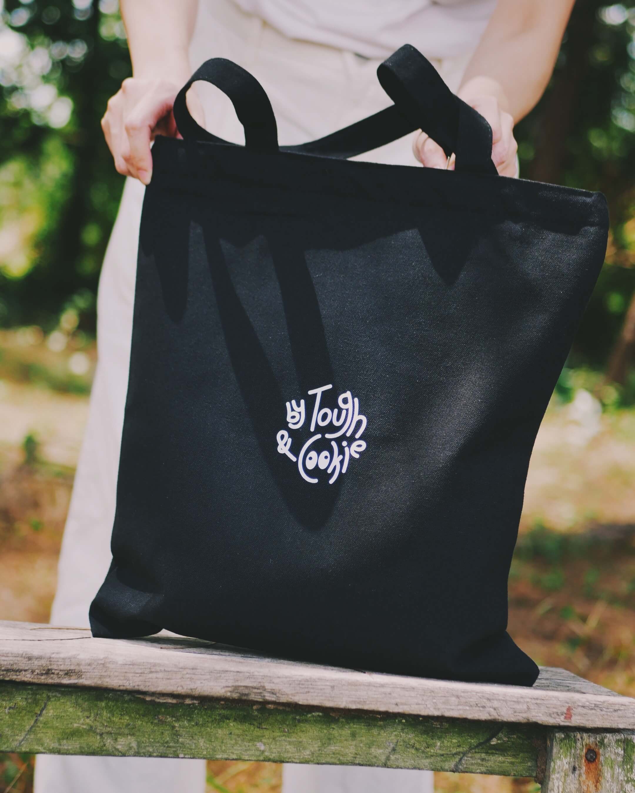 Black tote bag with a white logo held by a person outdoors