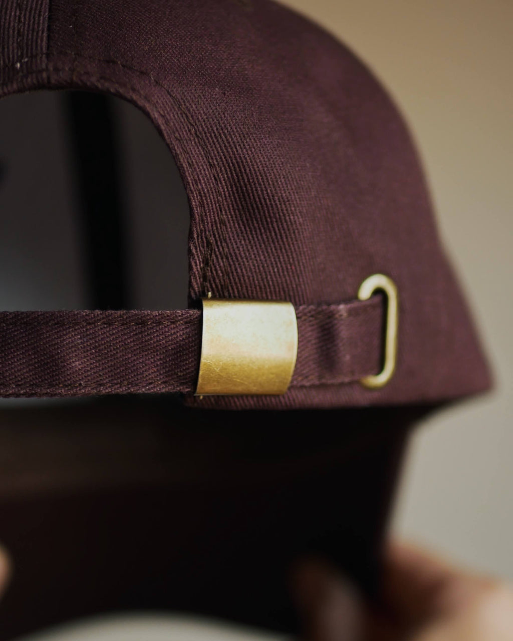 Close-up of the back strap of a dark brown baseball cap with a gold metal slider and adjustment buckle, held by a hand.
