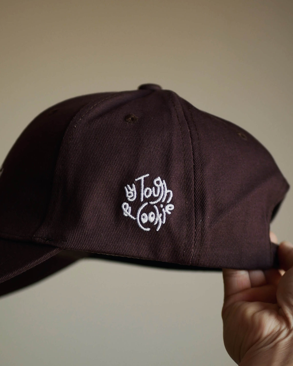 A dark brown baseball cap held by a hand, featuring white embroidered logo.