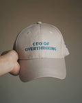 A hand holding a beige baseball cap with blue embroidered text reading 'CEO OF OVERTHINKING' against a plain taupe background.