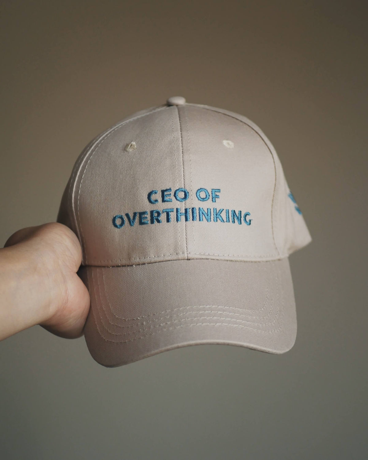 A hand holding a beige baseball cap with blue embroidered text reading 'CEO OF OVERTHINKING' against a plain taupe background.