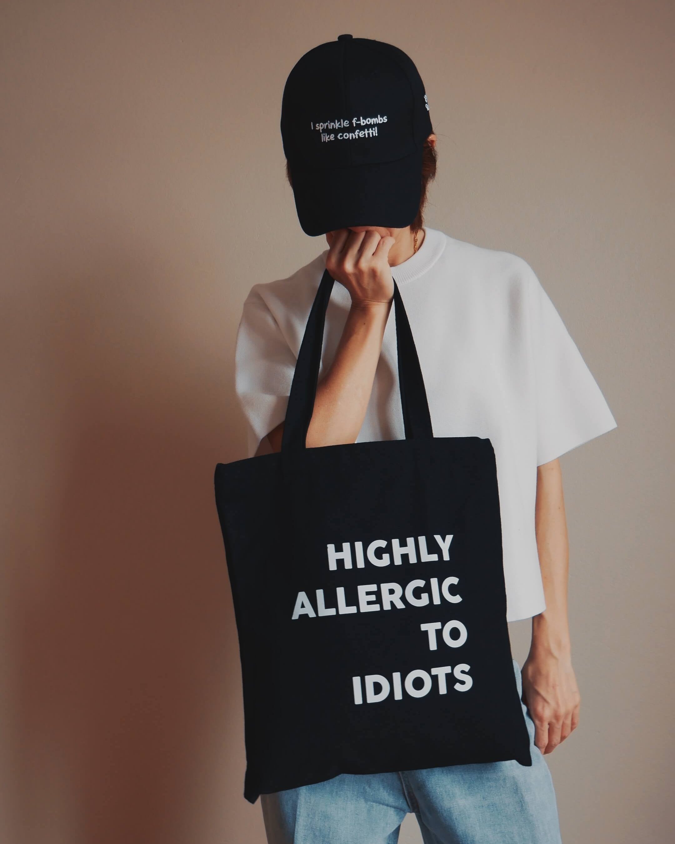 Person holding a black tote bag with text, wearing a black cap against a plain background