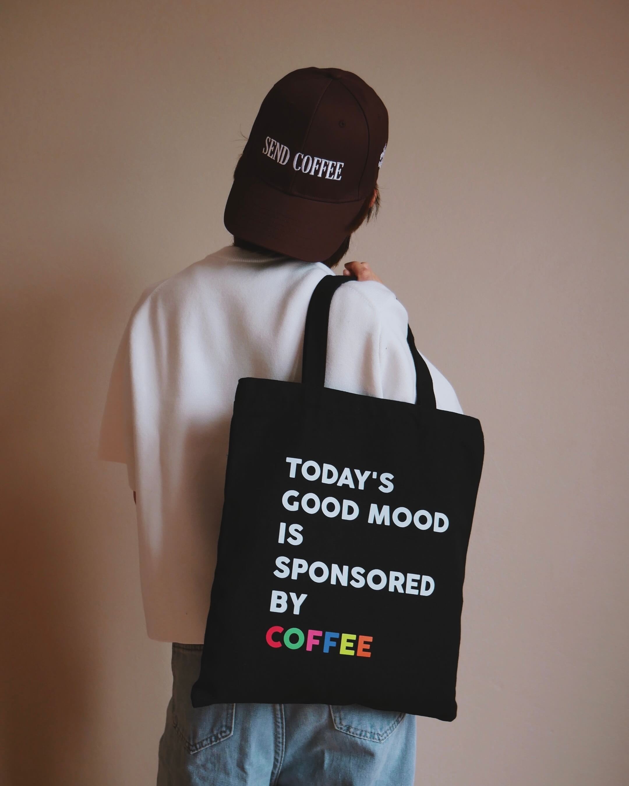 Person wearing a cap and holding a tote bag with a coffee-themed message.