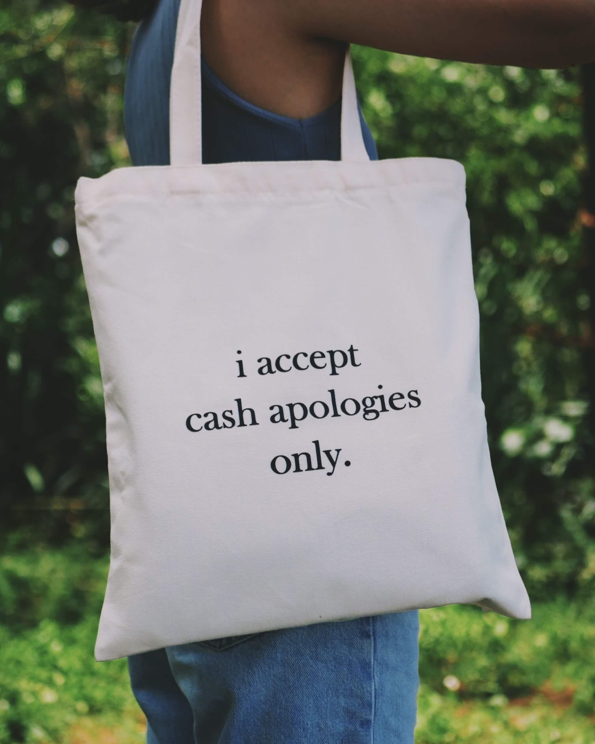 Tote bag with 'i accept cash apologies only.' text held by a person outdoors.