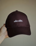 A hand holding a dark brown baseball cap with 'coffee addict' embroidered in white on the front, set against a plain beige background.