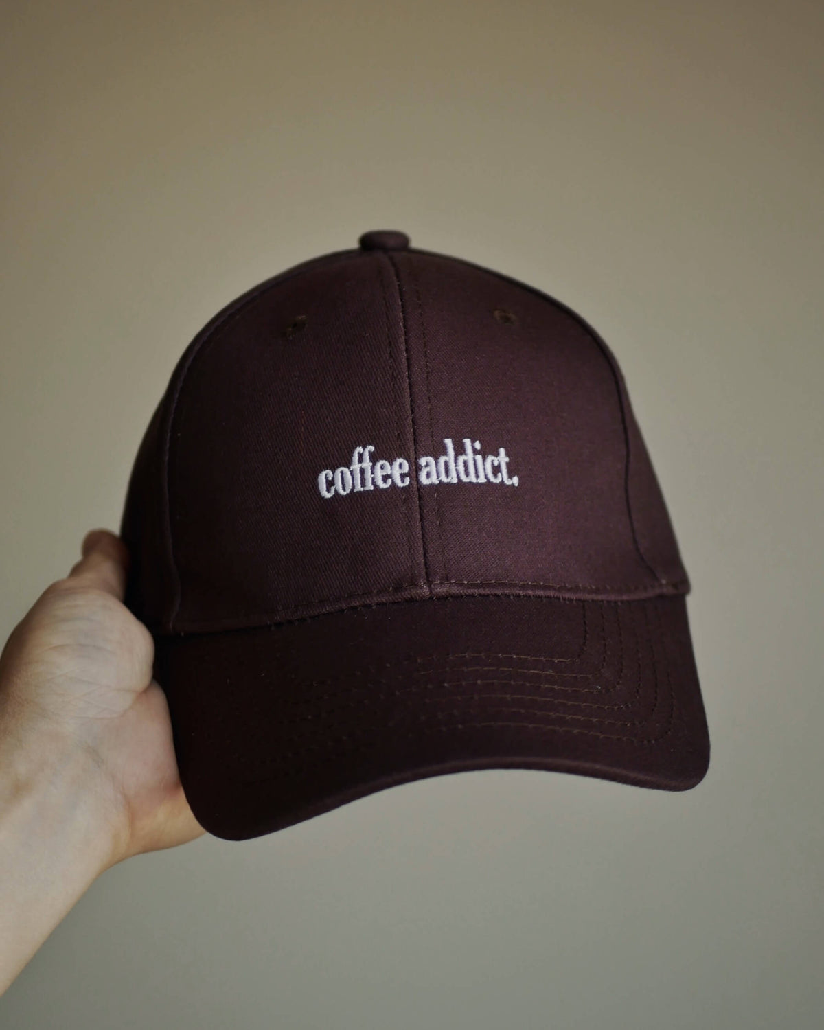A hand holding a dark brown baseball cap with 'coffee addict' embroidered in white on the front, set against a plain beige background.