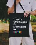 Person holding a black tote bag with text 'Today's good mood is sponsored by coffee' in an outdoor setting.