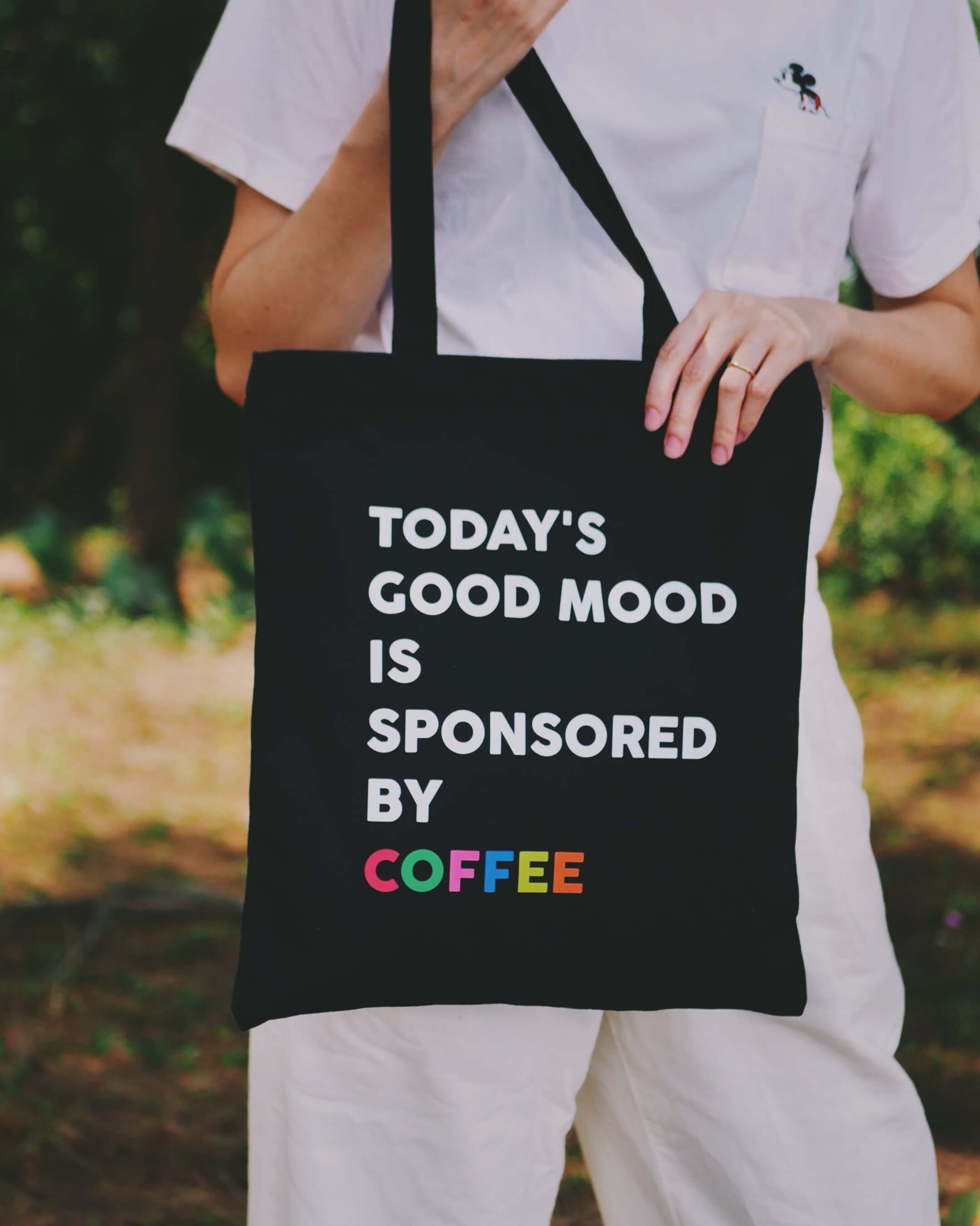 Person holding a black tote bag with text 'Today's good mood is sponsored by coffee' in an outdoor setting.