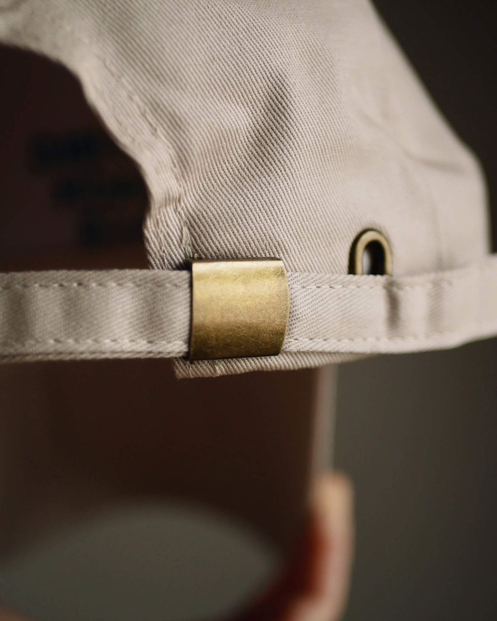 Close-up of a light beige canvas baseball cap with a brass metal buckle strap adjustment, held up by a hand.