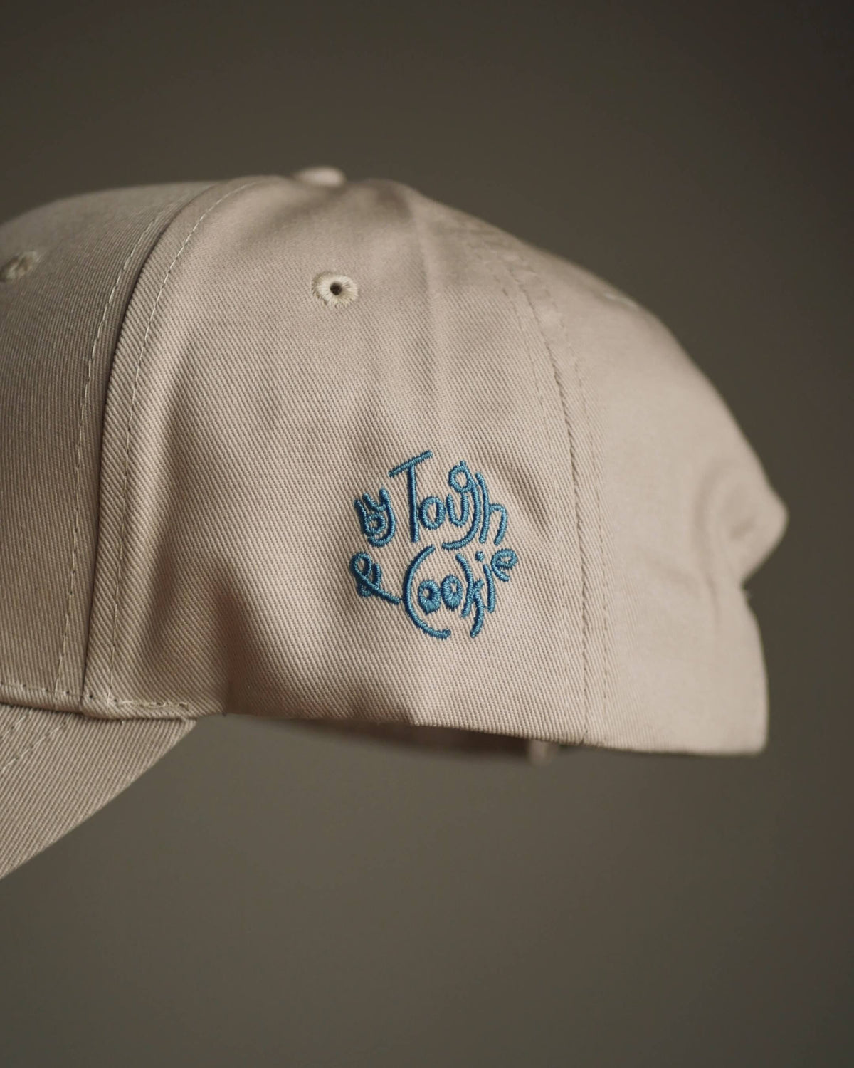 A close-up of a light beige baseball cap with blue embroidered logo on the side, set against a dark gray background.