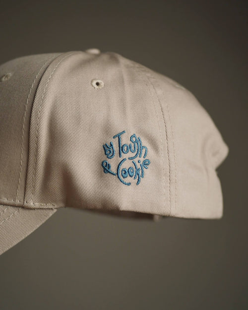 A close-up of a light beige baseball cap with blue embroidered logo on the side, set against a dark gray background.