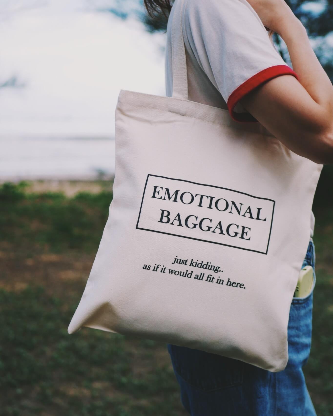 Tote bag with 'EMOTIONAL BAGGAGE' text held by a person outdoors.