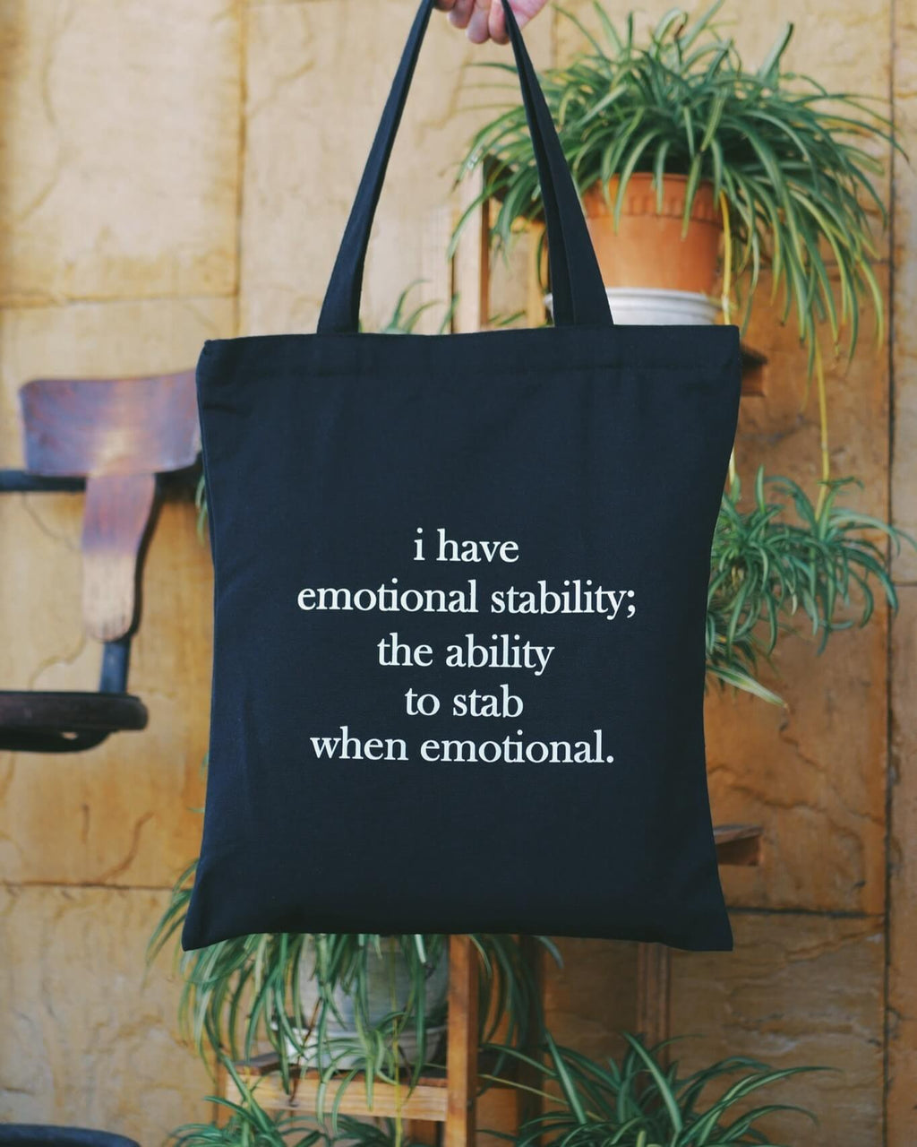 Black tote bag with humorous text about emotional stability held by a person in front of a plant.