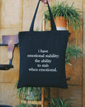 Black tote bag with humorous text about emotional stability held by a person in front of a plant.