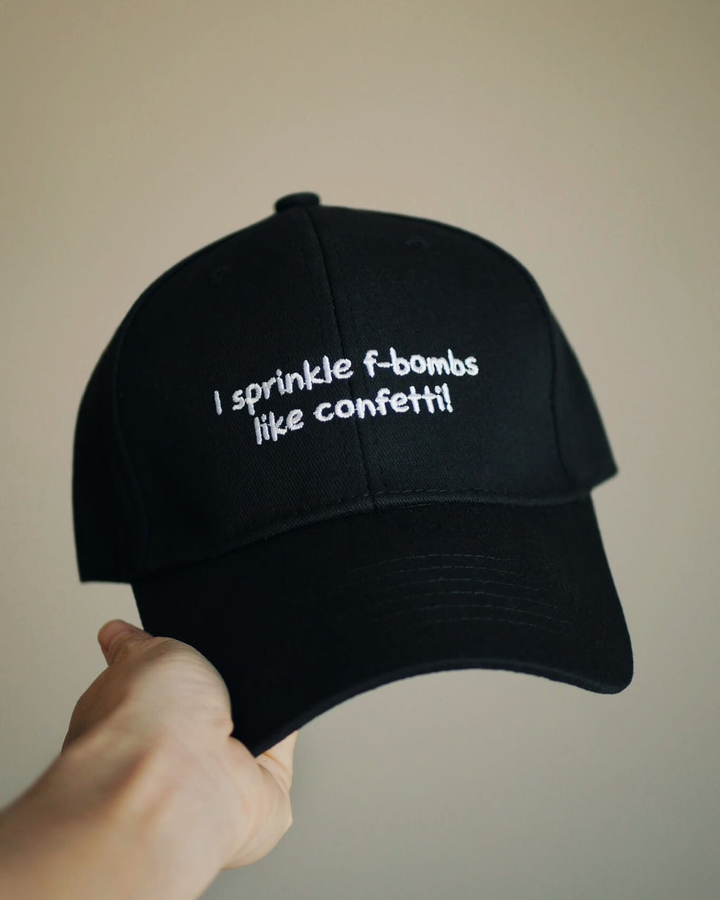 A black baseball cap held by a hand, featuring white embroidered text that reads 'I sprinkle f-bombs like confetti!' against a plain beige background.