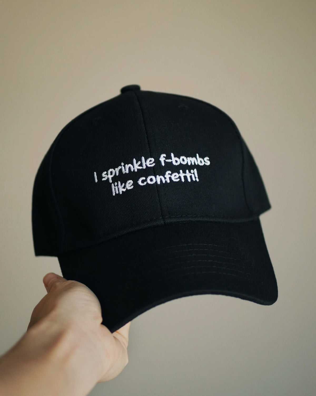 A black baseball cap held by a hand, featuring white embroidered text that reads 'I sprinkle f-bombs like confetti!' against a plain beige background.