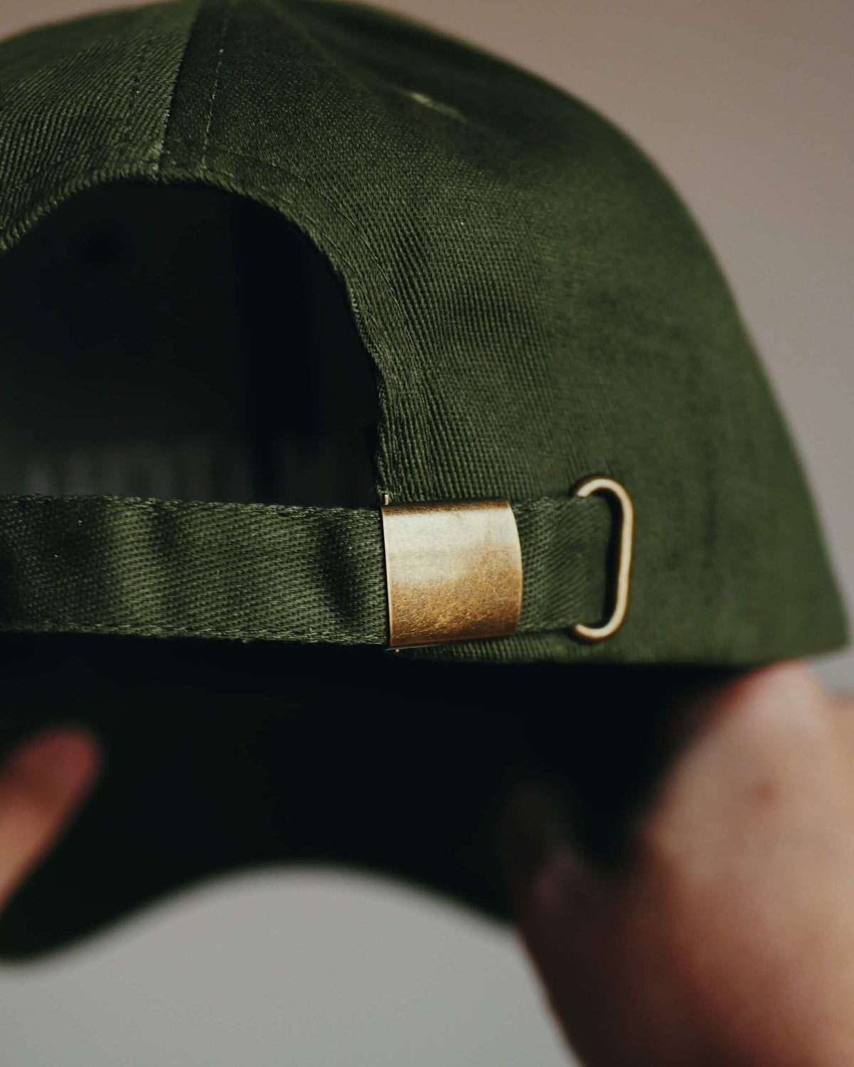 Close-up of the back of an olive green baseball cap being held by hands, showing a brass-colored metal slider buckle on the adjustable strap.