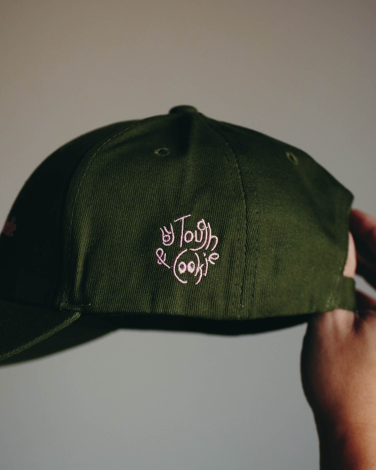 An olive green baseball cap held by a hand, featuring pink embroidered logo.