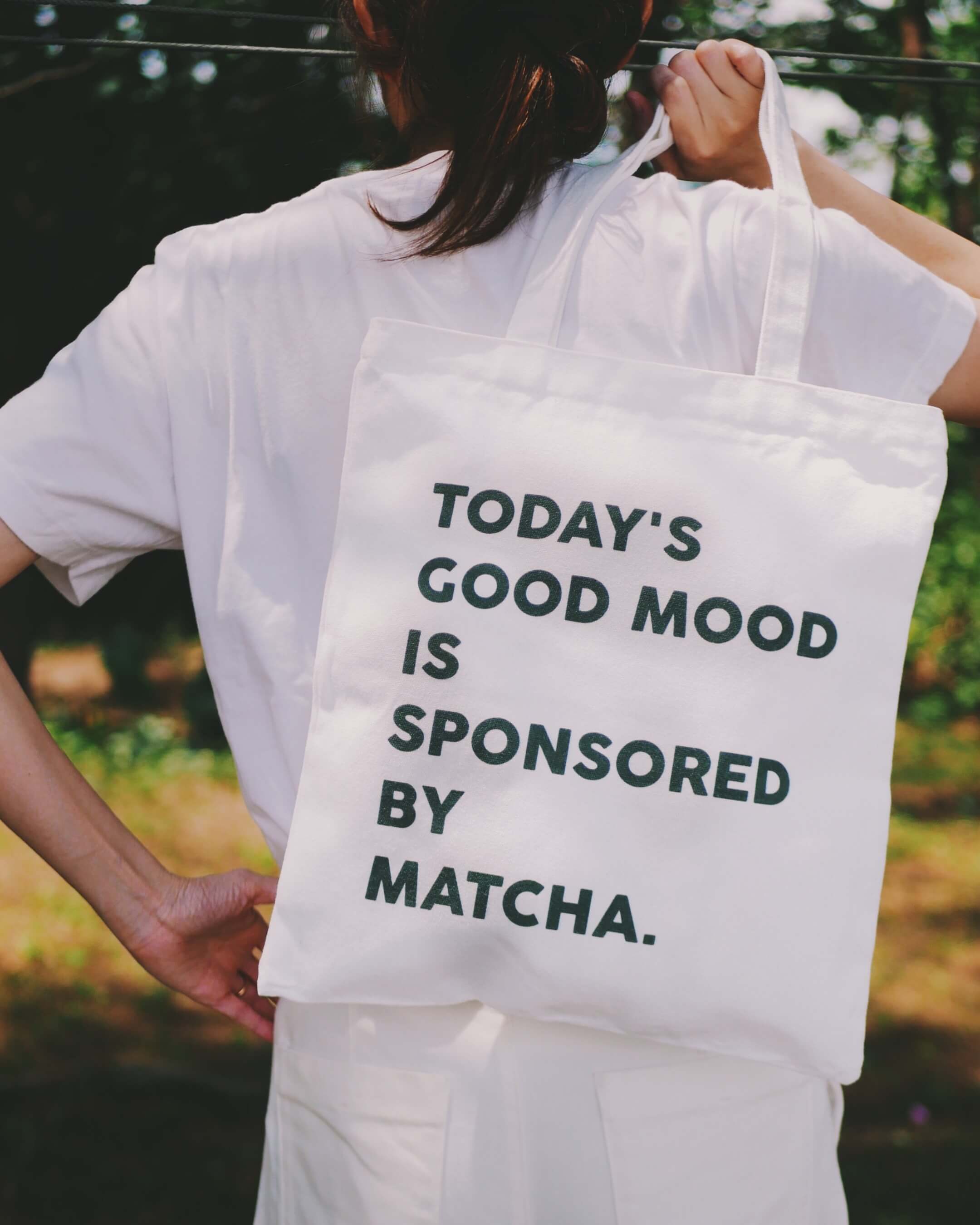 Person holding a white tote bag with text 'Today's good mood is sponsored by matcha' against a blurred outdoor background.