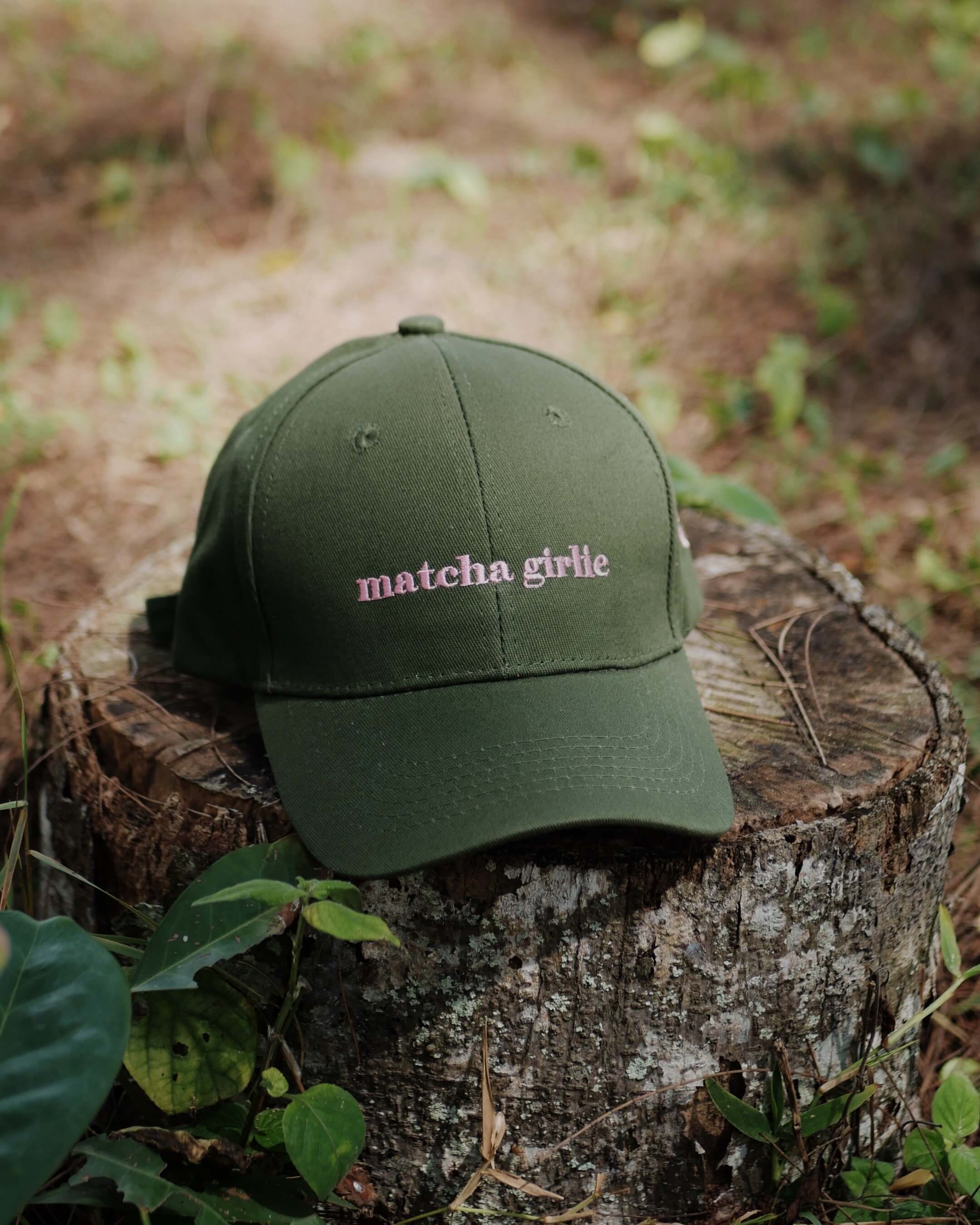 Green cap with 'matcha girlie' text on a wooden stump in a natural setting