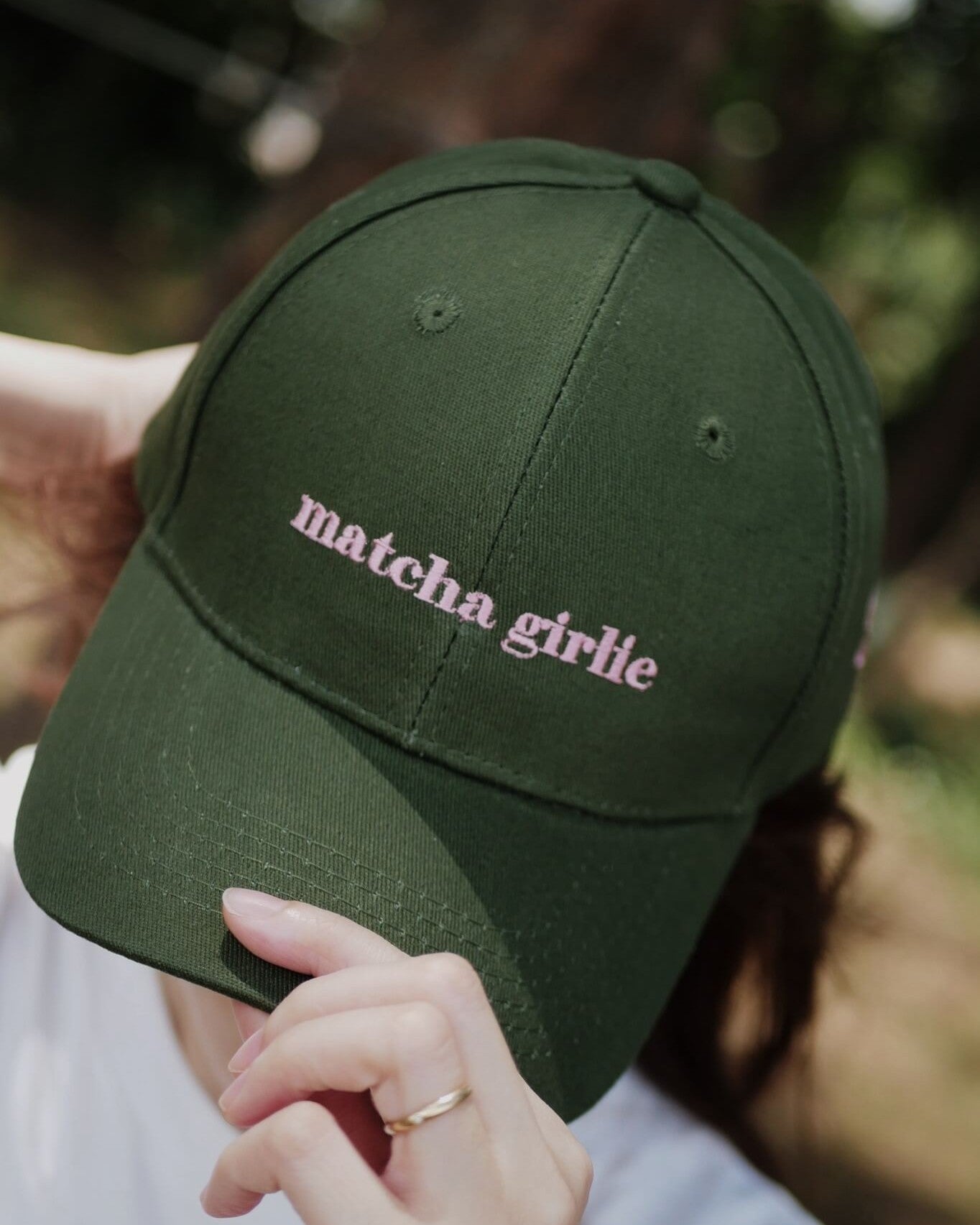Green cap with 'matcha girlie' text held by a person against a blurred natural background