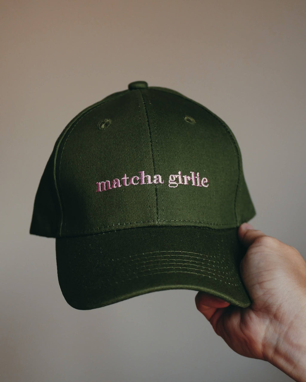 An olive green baseball cap with 'matcha girlie' embroidered in pink on the front, held by a hand against a plain beige background.