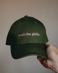 An olive green baseball cap with 'matcha girlie' embroidered in pink on the front, held by a hand against a plain beige background.