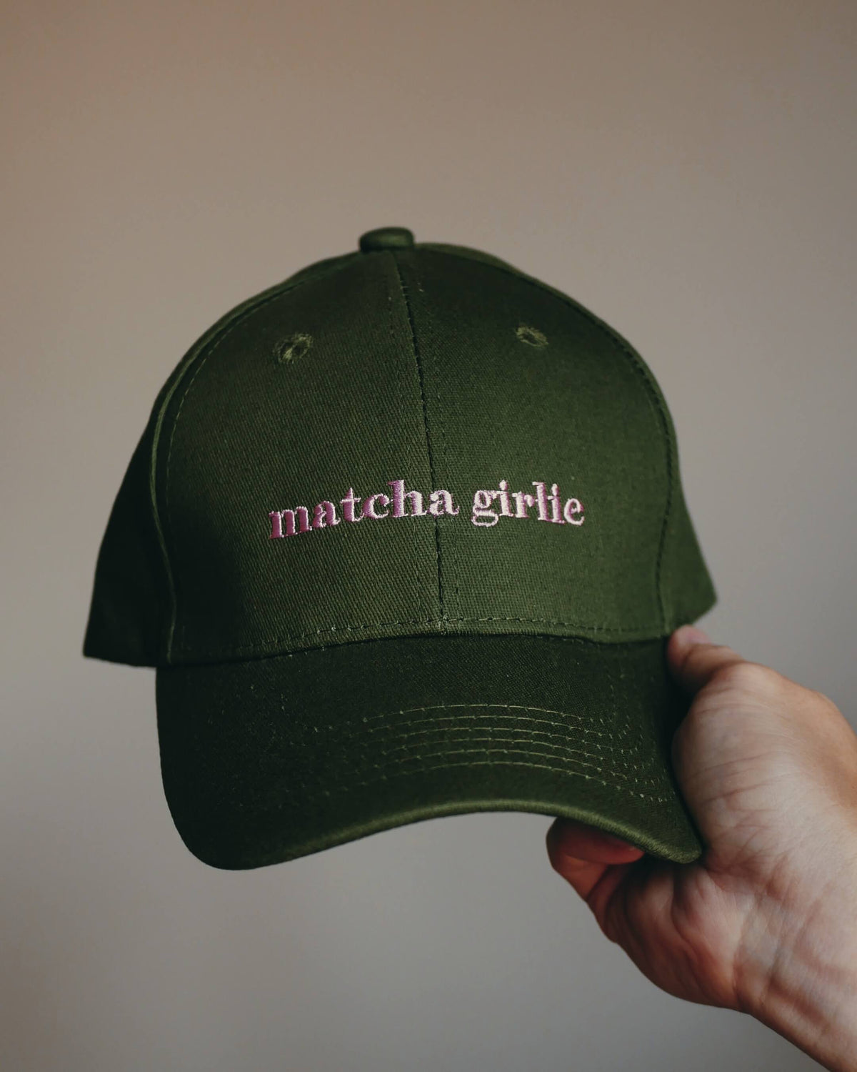 An olive green baseball cap with 'matcha girlie' embroidered in pink on the front, held by a hand against a plain beige background.