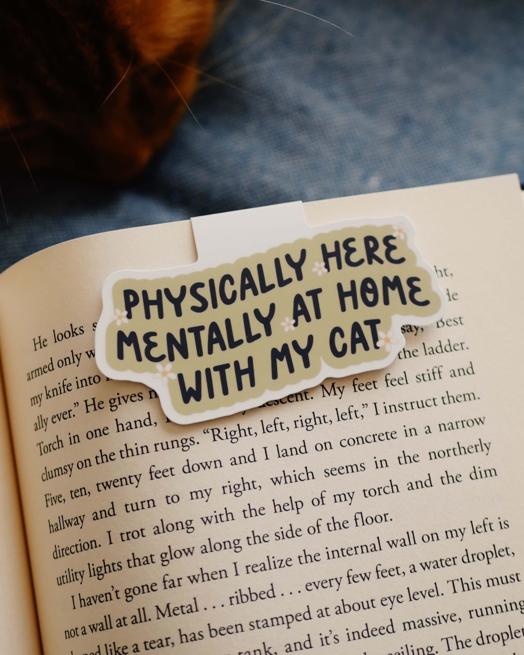 Magnetic bookmark with 'Physically here, mentally with my cat' design clipped on an open book.