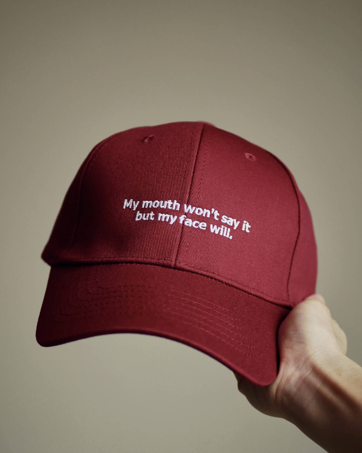 A maroon baseball cap held by a hand, featuring white embroidered text that reads 'My mouth won't say it but my face will.' against a solid beige background.