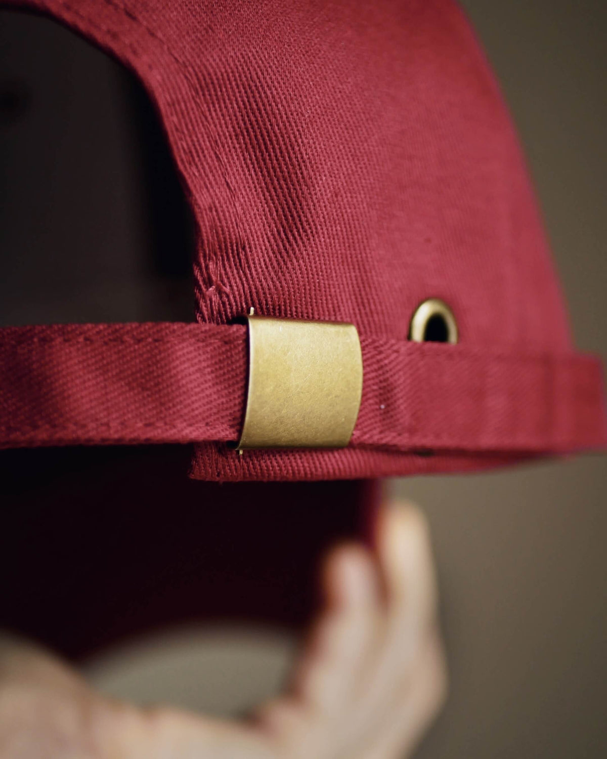 Close-up of a burgundy baseball cap with a gold metal buckle and grommet, held by a hand.