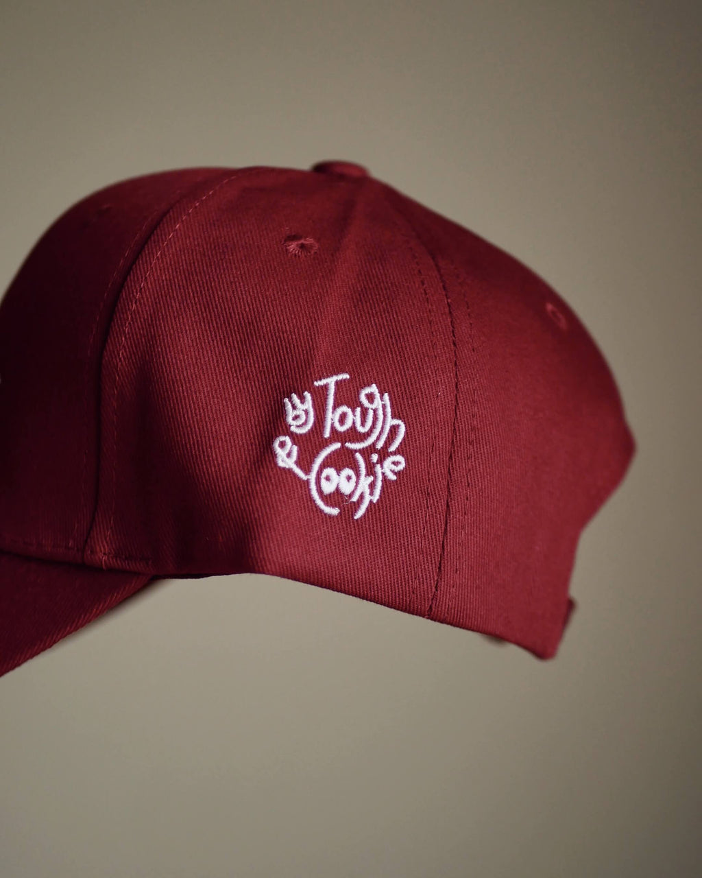 A close-up of a dark red baseball cap featuring white embroidered logo, set against a plain beige background.