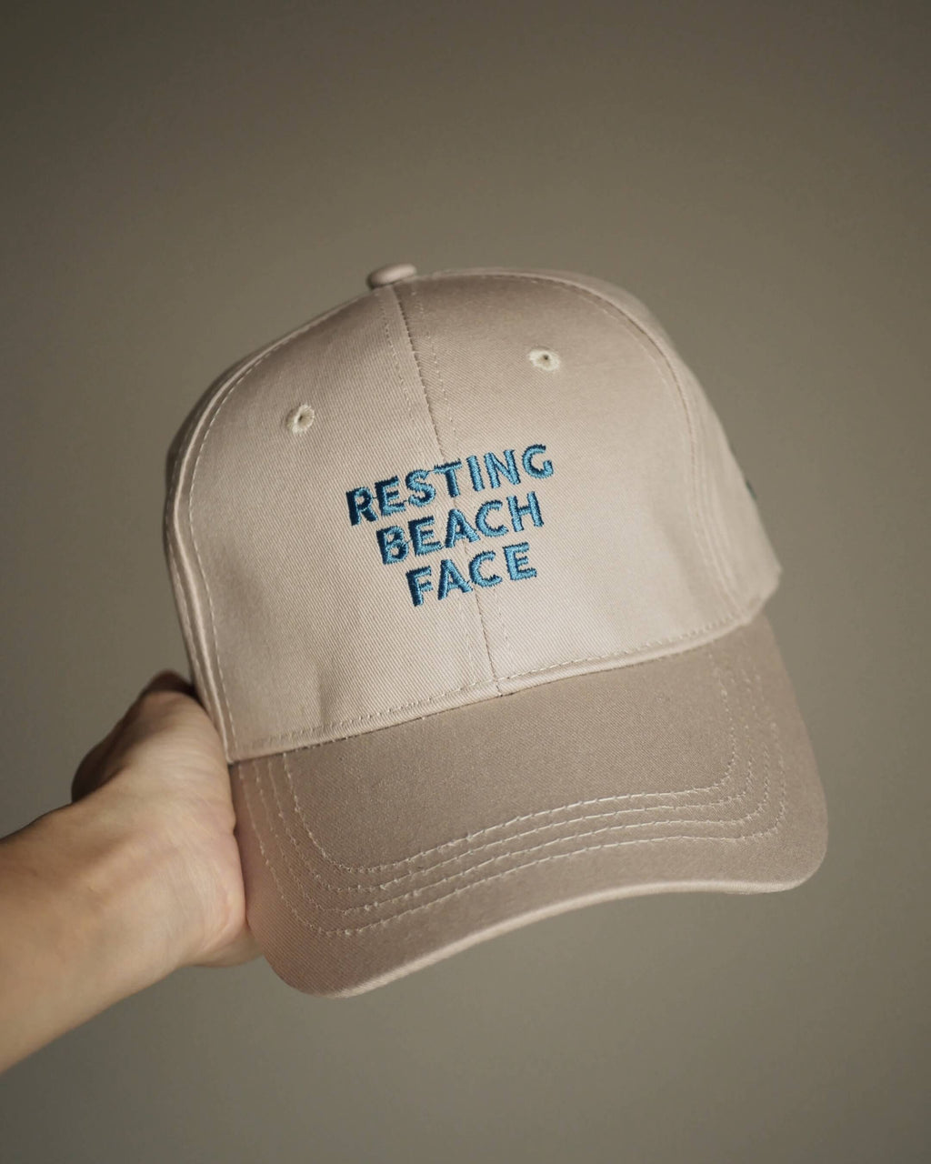 A beige baseball cap held by a hand, featuring blue embroidered text on the front that reads 'RESTING BEACH FACE'.