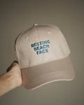 A beige baseball cap held by a hand, featuring blue embroidered text on the front that reads 'RESTING BEACH FACE'.