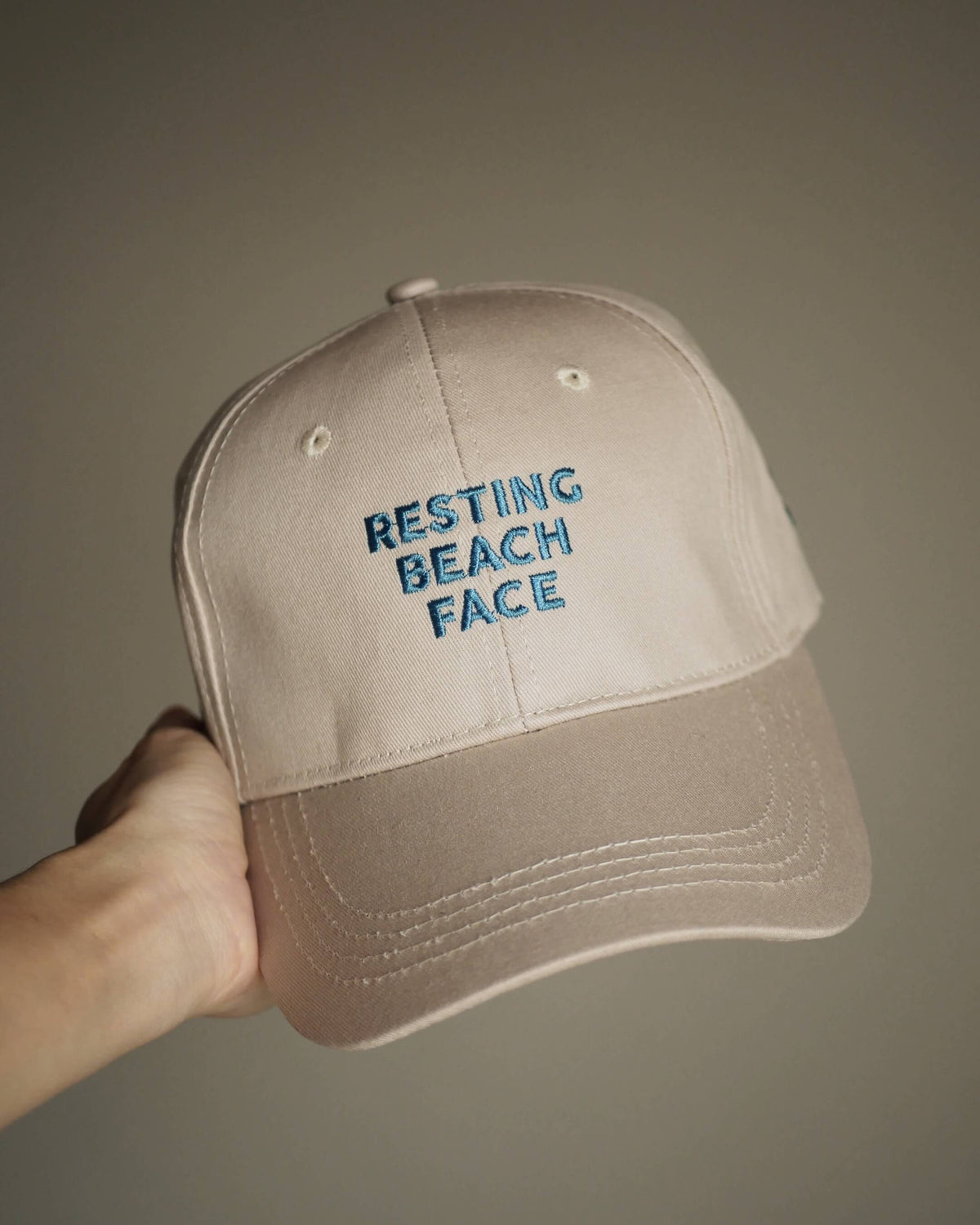 A beige baseball cap held by a hand, featuring blue embroidered text on the front that reads 'RESTING BEACH FACE'.