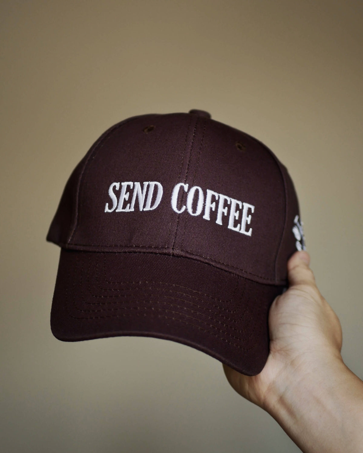 A dark brown baseball cap with 'SEND COFFEE' embroidered in white on the front, held by a hand against a plain beige background.