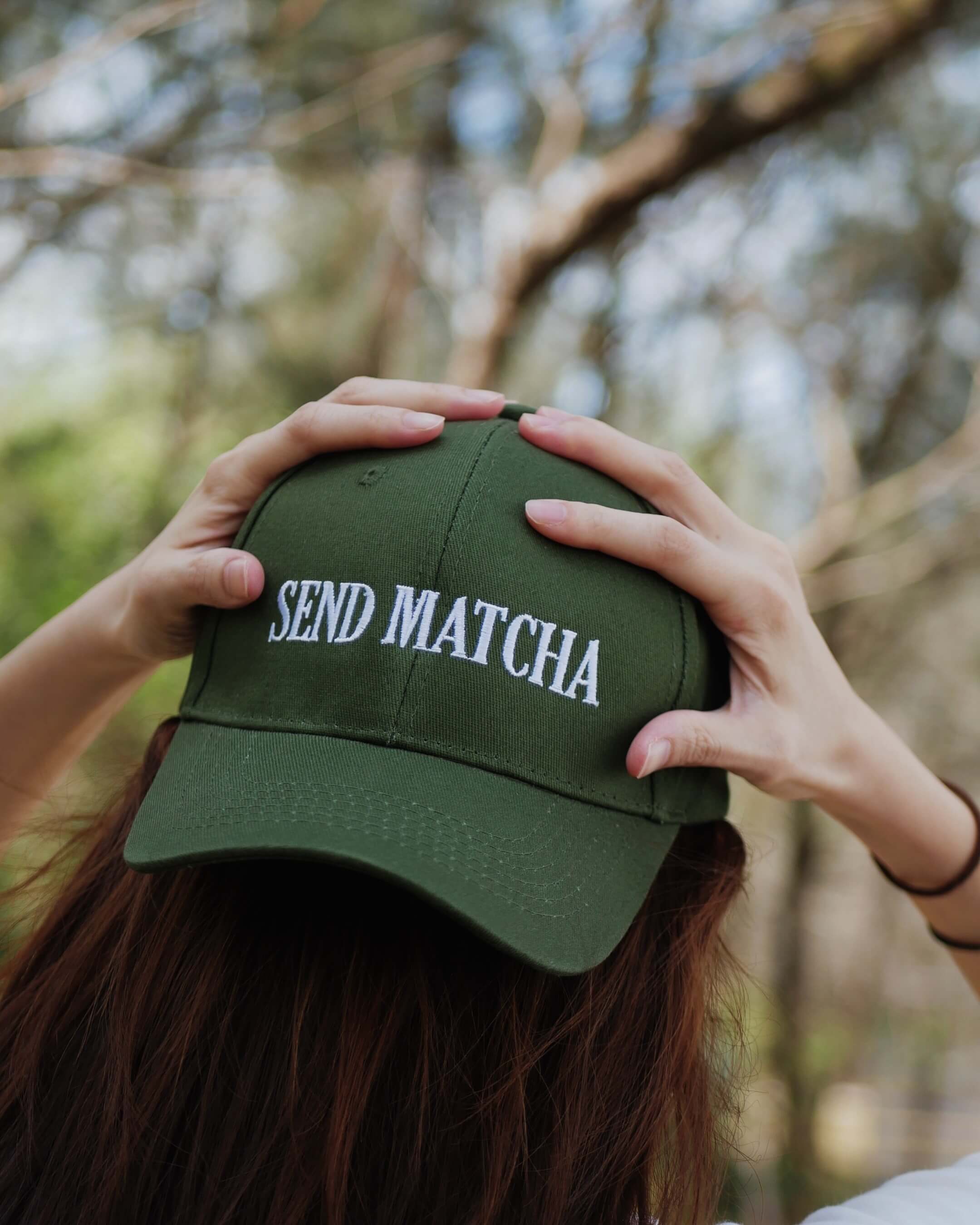 Person holding a green cap with 'SEND MATCHA' text against a blurred natural background