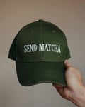 An olive green baseball cap with 'SEND MATCHA' embroidered in white on the front, held by a hand against a plain beige background.