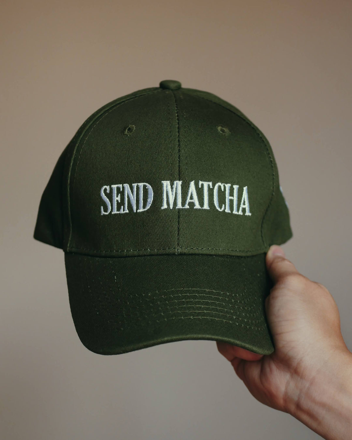 An olive green baseball cap with 'SEND MATCHA' embroidered in white on the front, held by a hand against a plain beige background.