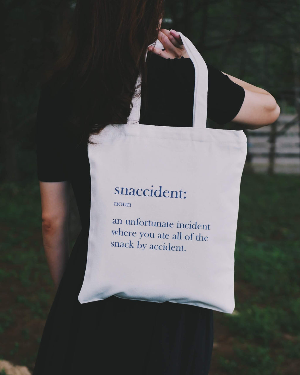 A person holding a 'Snaccident' tote bag outdoor