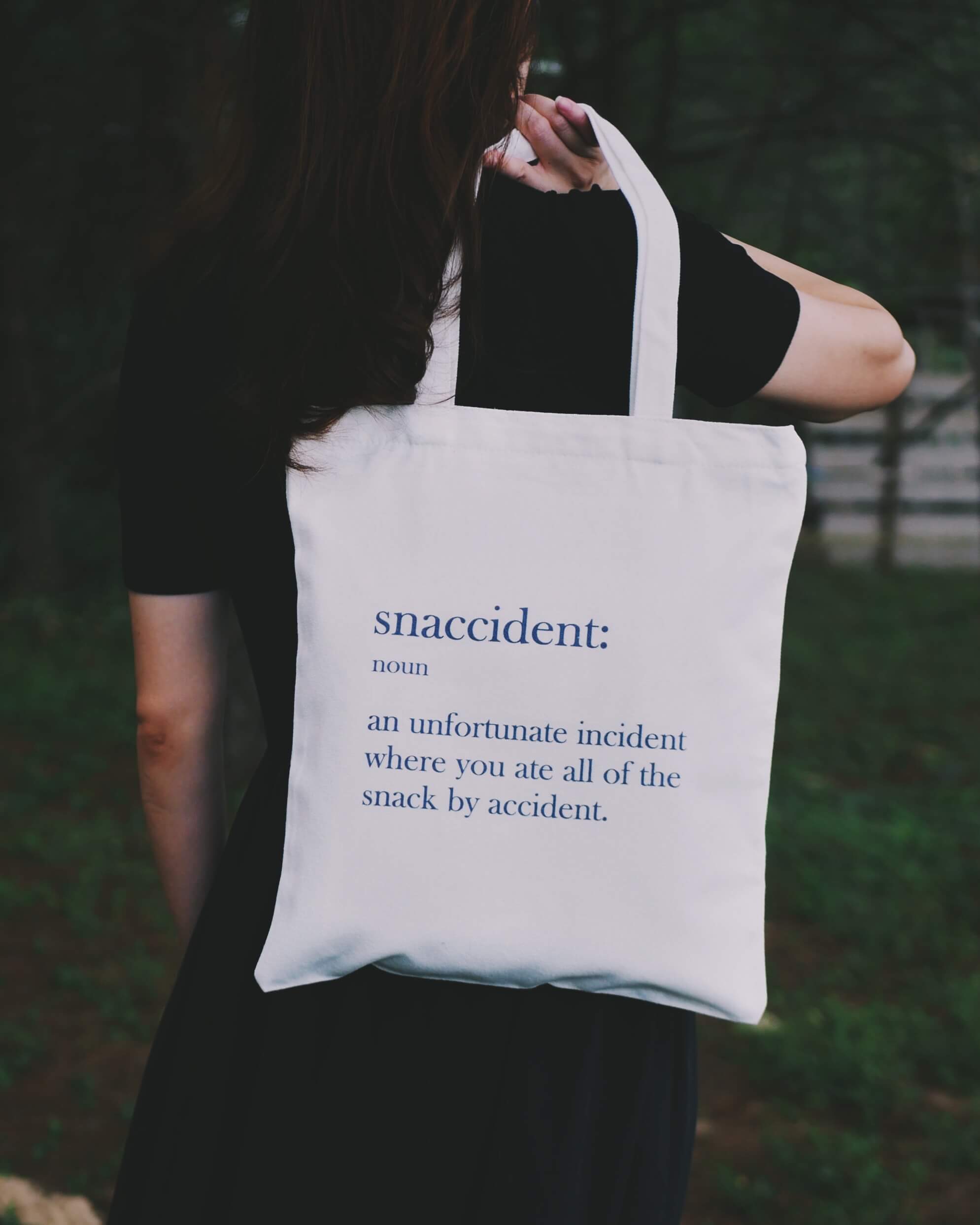 A person holding a 'Snaccident' tote bag outdoor
