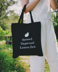 Person holding a black tote bag with "stressed depressed lemon zest" text outdoors