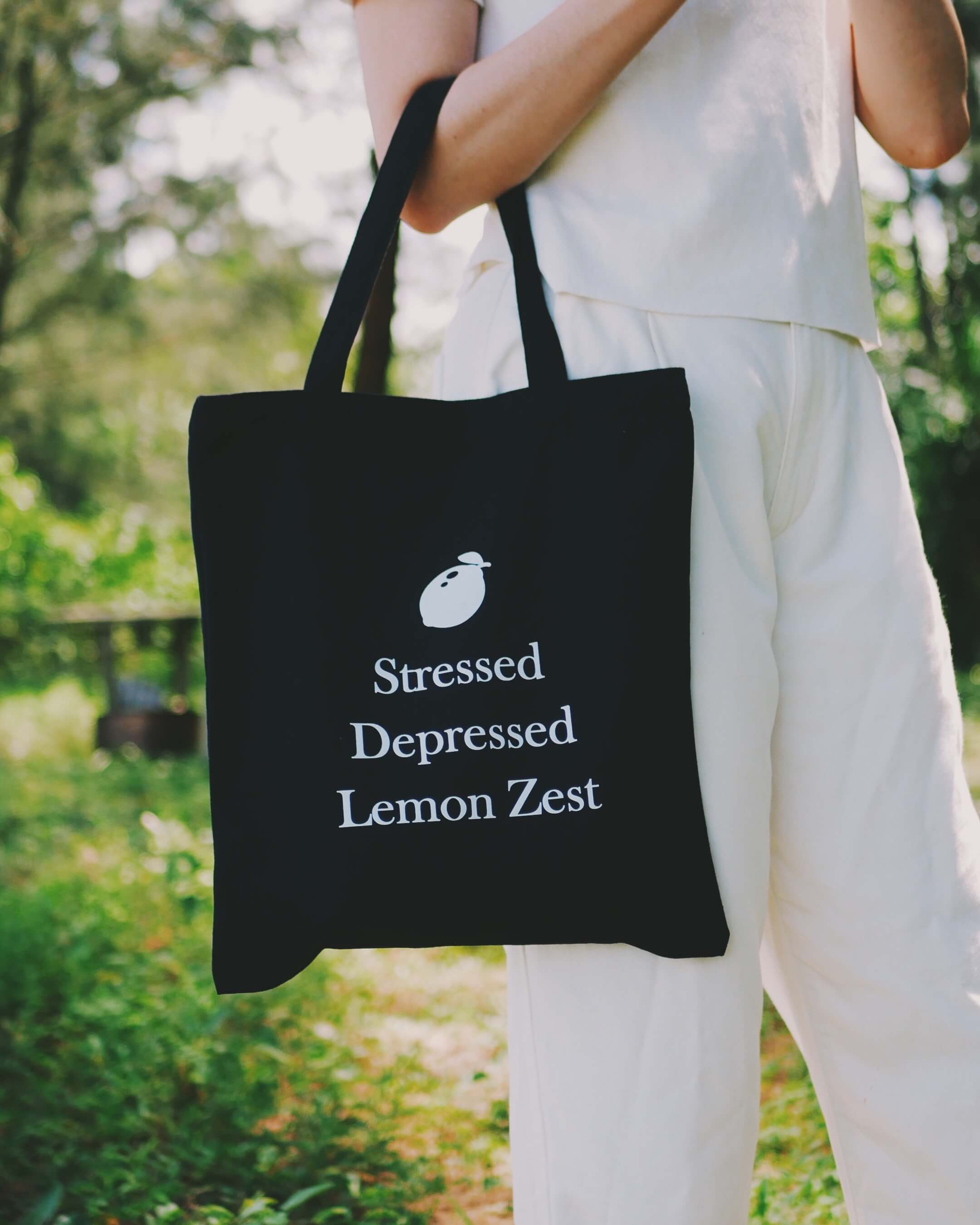 Person holding a black tote bag with "stressed depressed lemon zest" text outdoors