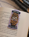 Magnetic bookmark with "Turn Pages With Caution" design clipped on an open book.
