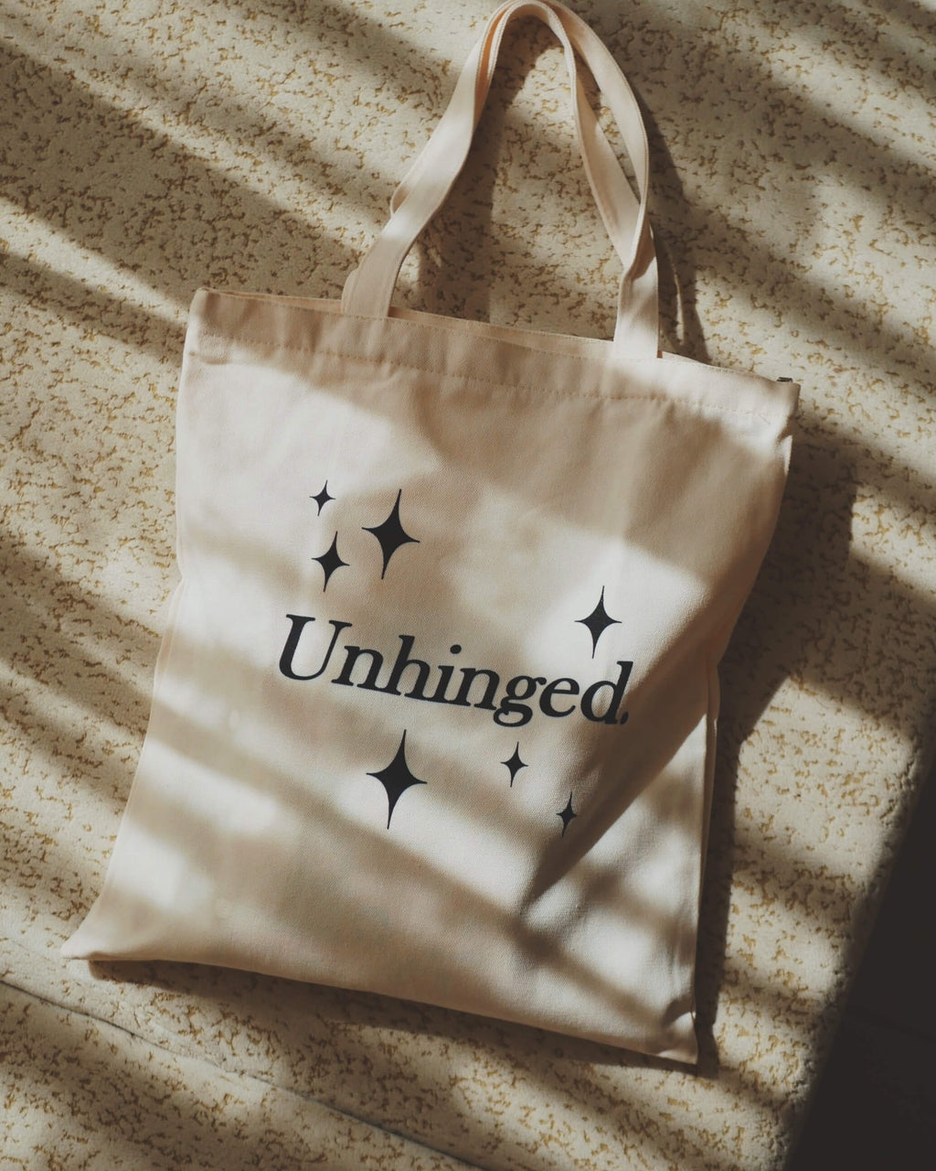 Beige tote bag with 'Unhinged' text and star designs on a textured surface