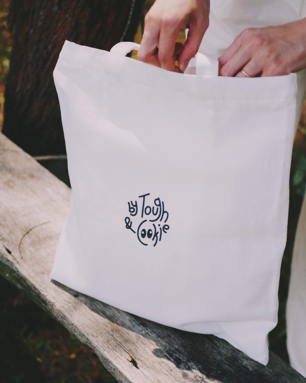 White tote bag with 'By Tough & Cookie' logo on a wooden surface