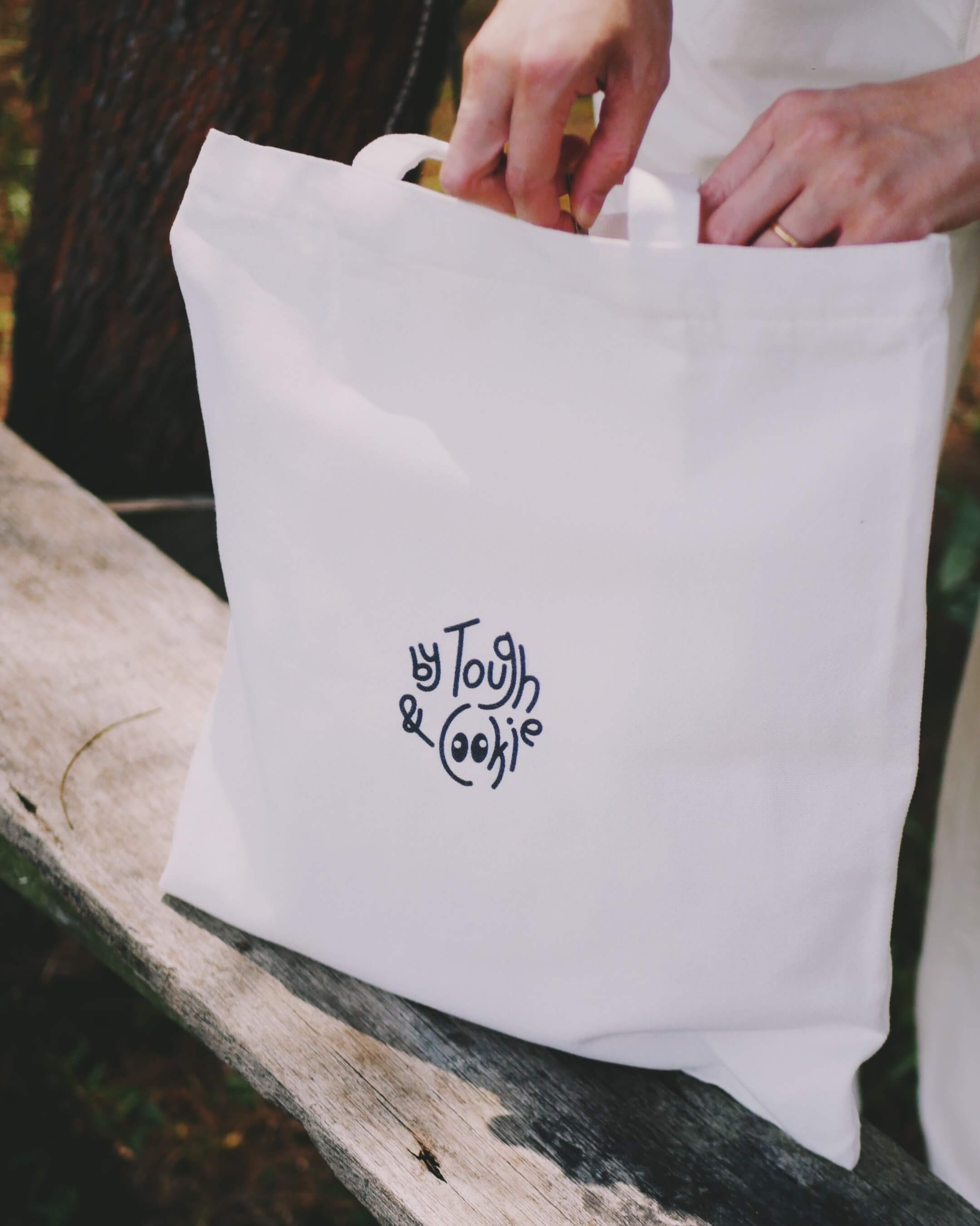White tote bag with 'By Tough & Cookie' logo on a wooden surface