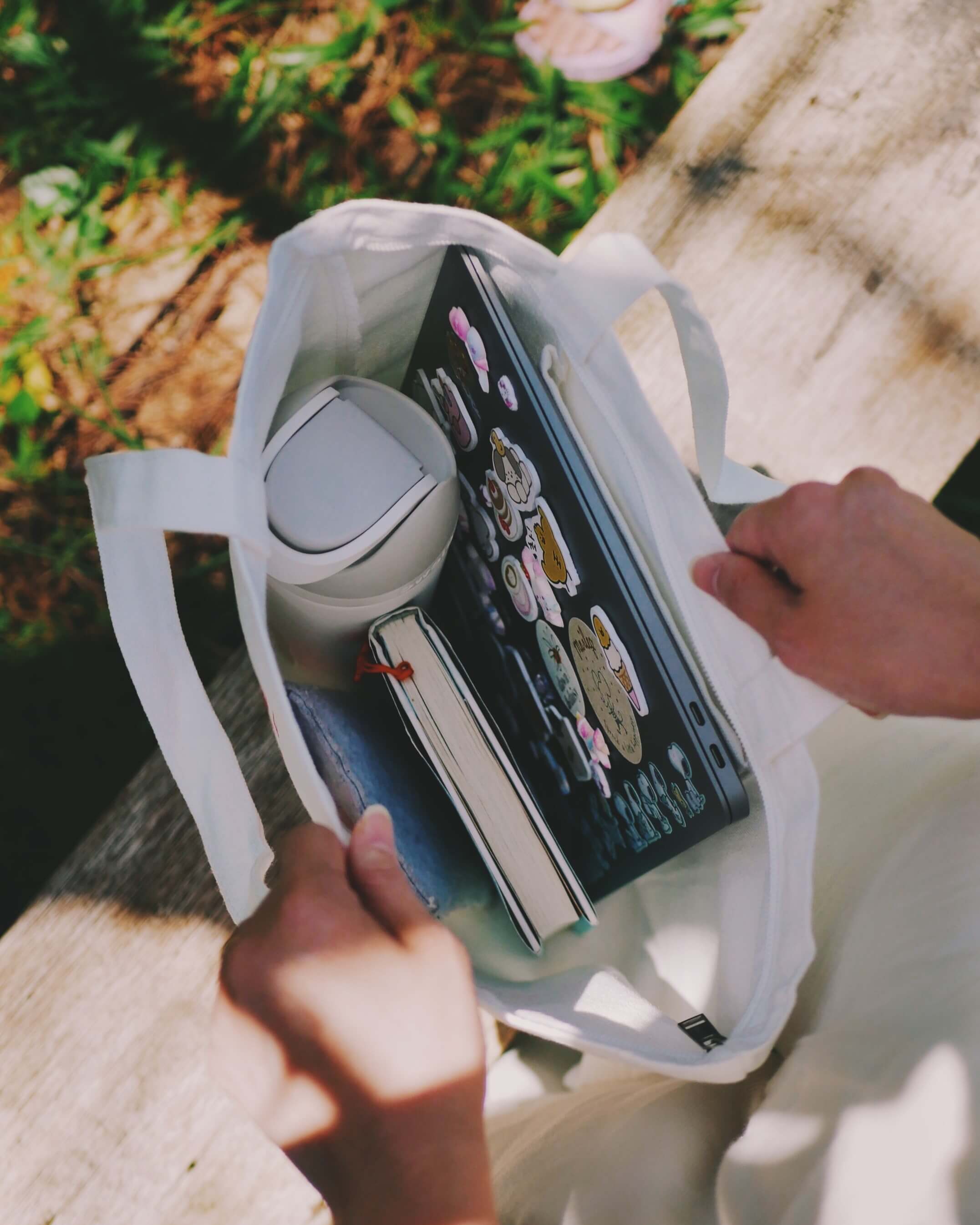 Person holding a white tote bag with a thermos, a book and a laptop on a wooden bench
